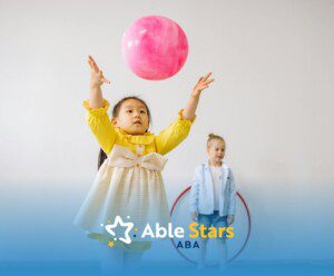 An autistic girl in yellow dress tossing a pink ball as another child watches at a playroom in MD.