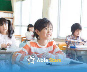 Smiling child in a striped shirt sits at a classroom desk, engaged and attentive. Other children study nearby. Bright, cheerful atmosphere.