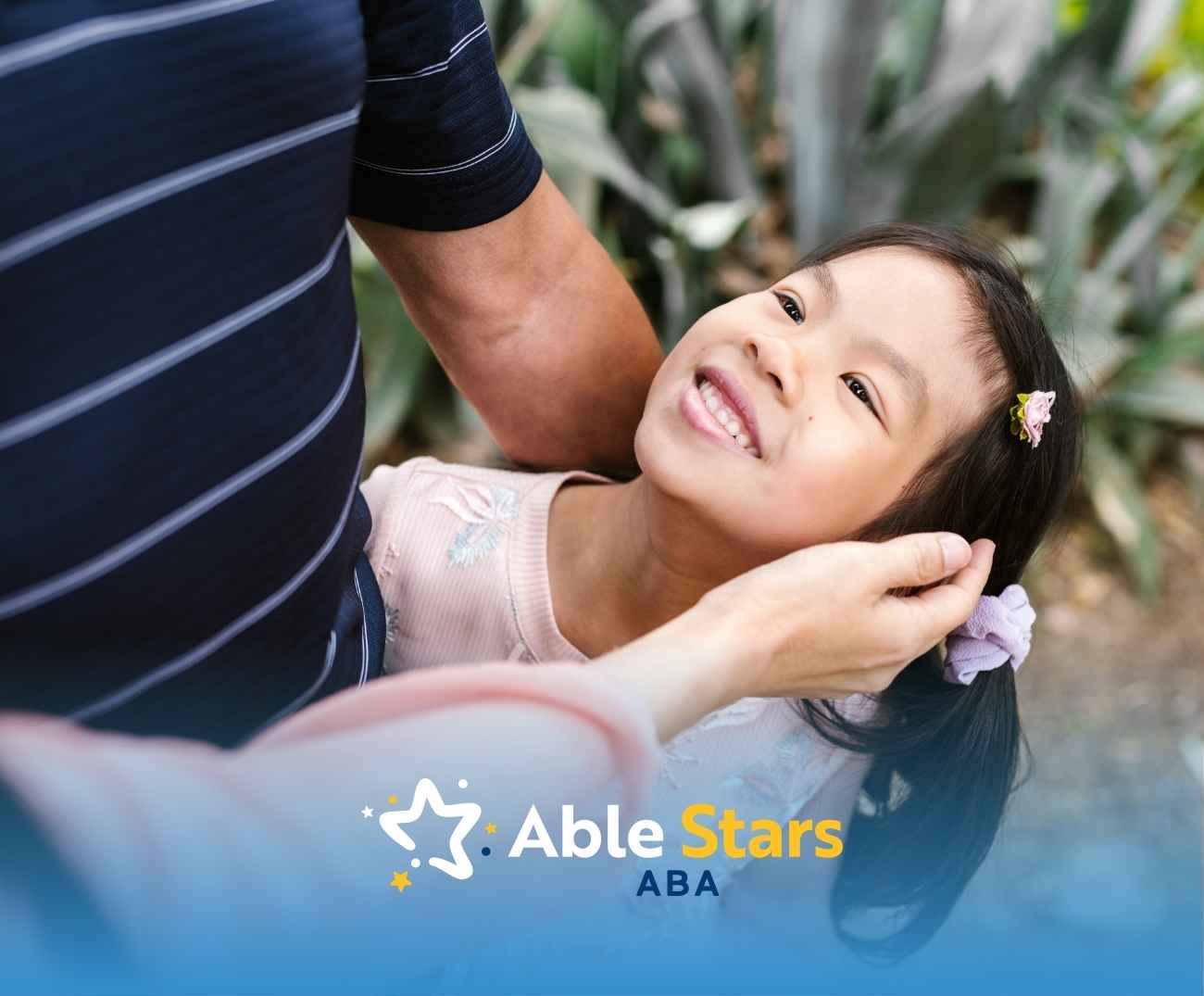 A young girl smiles joyfully, looking up as an adult gently touches her face. Lush greenery is blurred in the background.