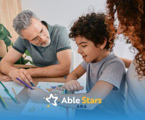 A smiling boy draws with colored pencils at a table, assisted by two adults. The scene conveys warmth and support.