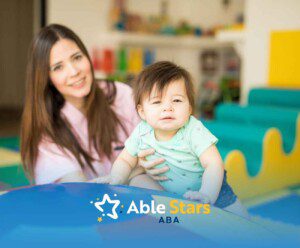 A smiling woman sits with a toddler in an indoor play area, featuring bright, colorful shapes.