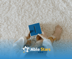 An autistic child sitting on soft carpet, holding a blue toy, looking down during ABA therapy session in Maryland.
