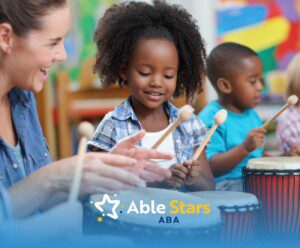 A smiling teacher and two young children play drums with wooden sticks in a colorful classroom.