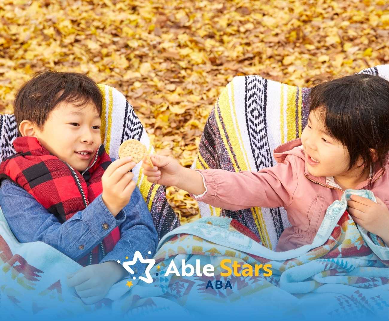 Two children sit on a blanket amidst autumn leaves, smiling and sharing a cookie. They are wrapped in colorful blankets.