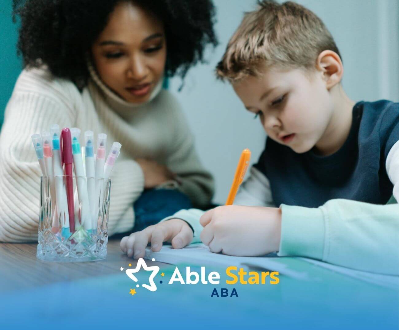 A young boy & ABA therapist sit together at a table, both focused & writing as they collaborate on a creative project.