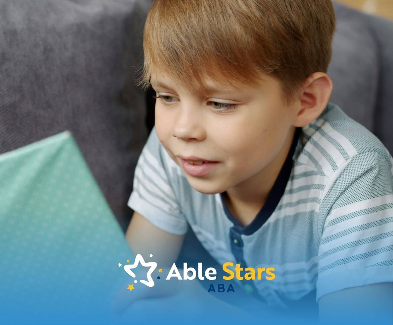 Child in striped shirt sits on gray couch, focused on reading a light blue book with subtle patterns.