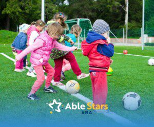 Children wearing jackets and hats stand in a row with soccer balls on a green turf field, ready to play.