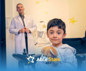 Child in a motorized wheelchair hugs a teddy bear while sitting near a doctor in a bright clinic setting.