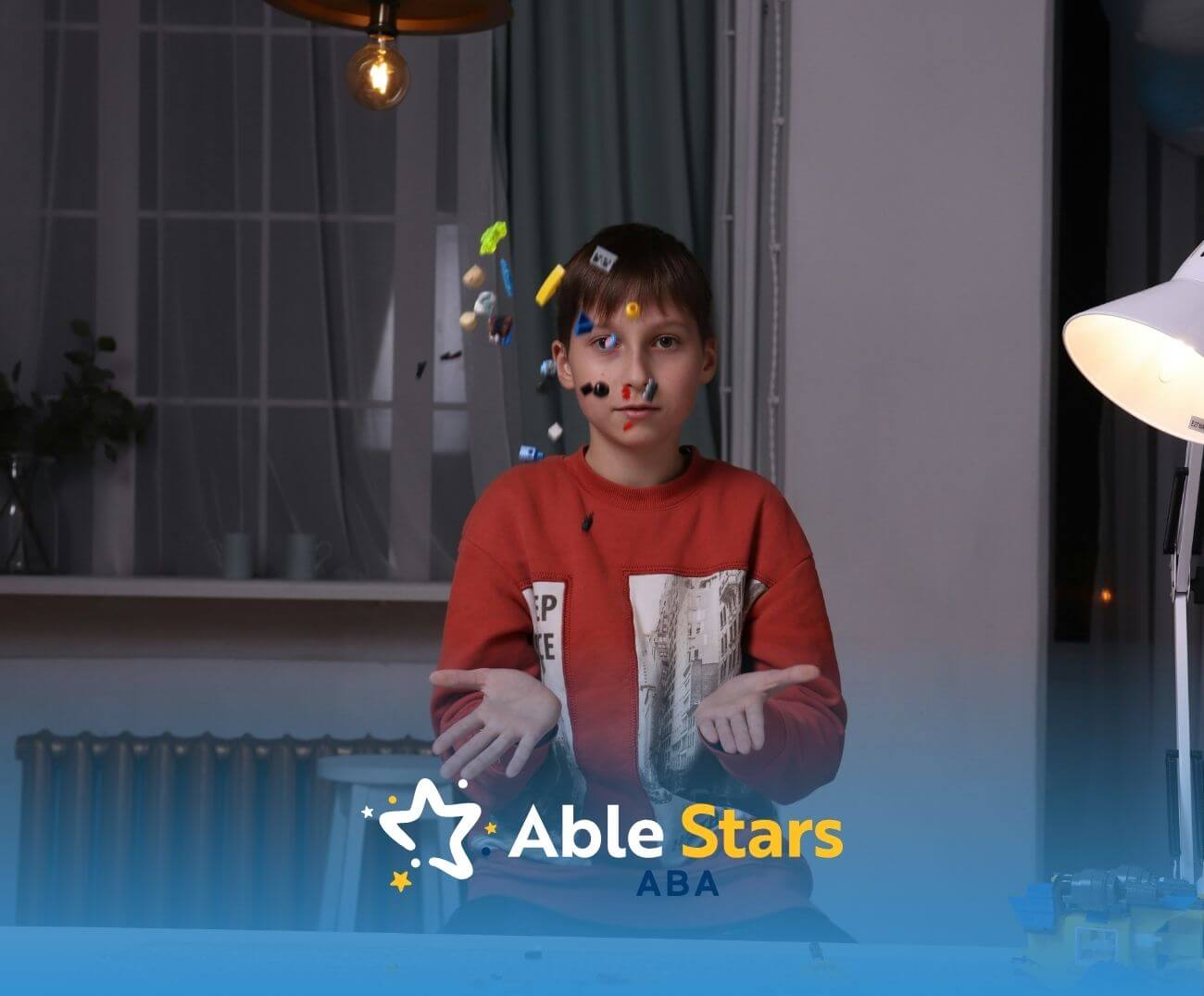 Child in red sweatshirt tosses LEGO pieces into air over table with scattered bricks and lit desk lamp nearby.