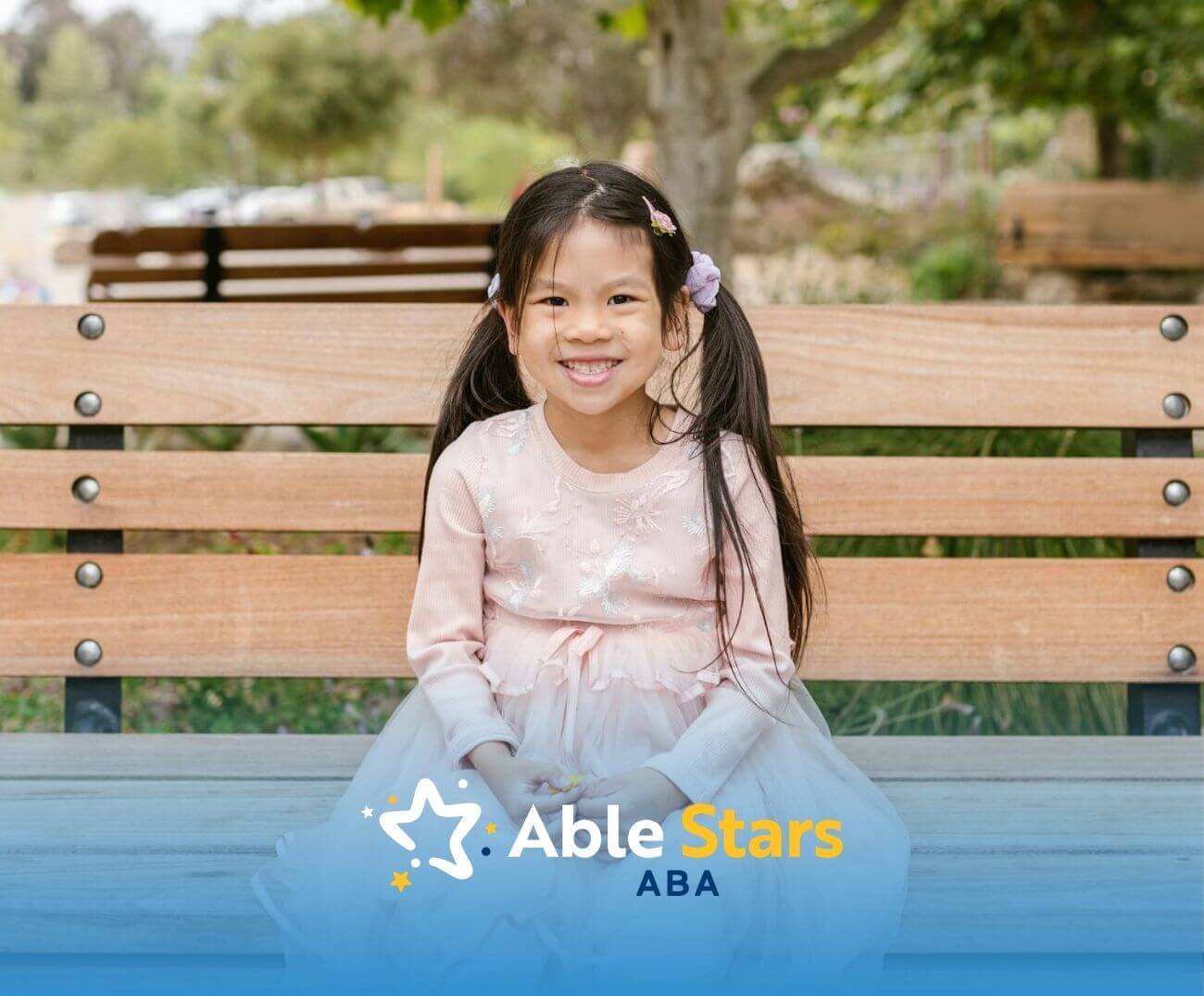 A girl in a pink dress smiles warmly while sitting on a park bench, enjoying the peaceful outdoor setting around her.