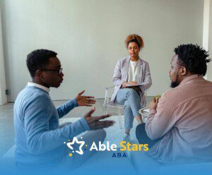 Three individuals converse in a sleek room, with one acting as mediator while the other two engage in discussion.