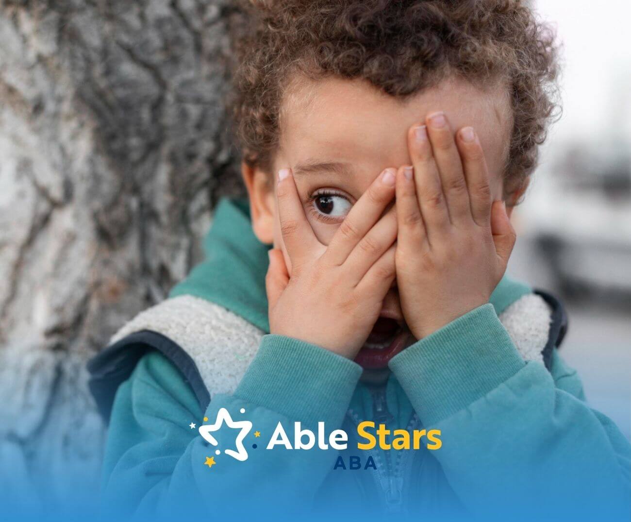 Boy with autism playing peek-a-boo by covering his face against a tree trunk.