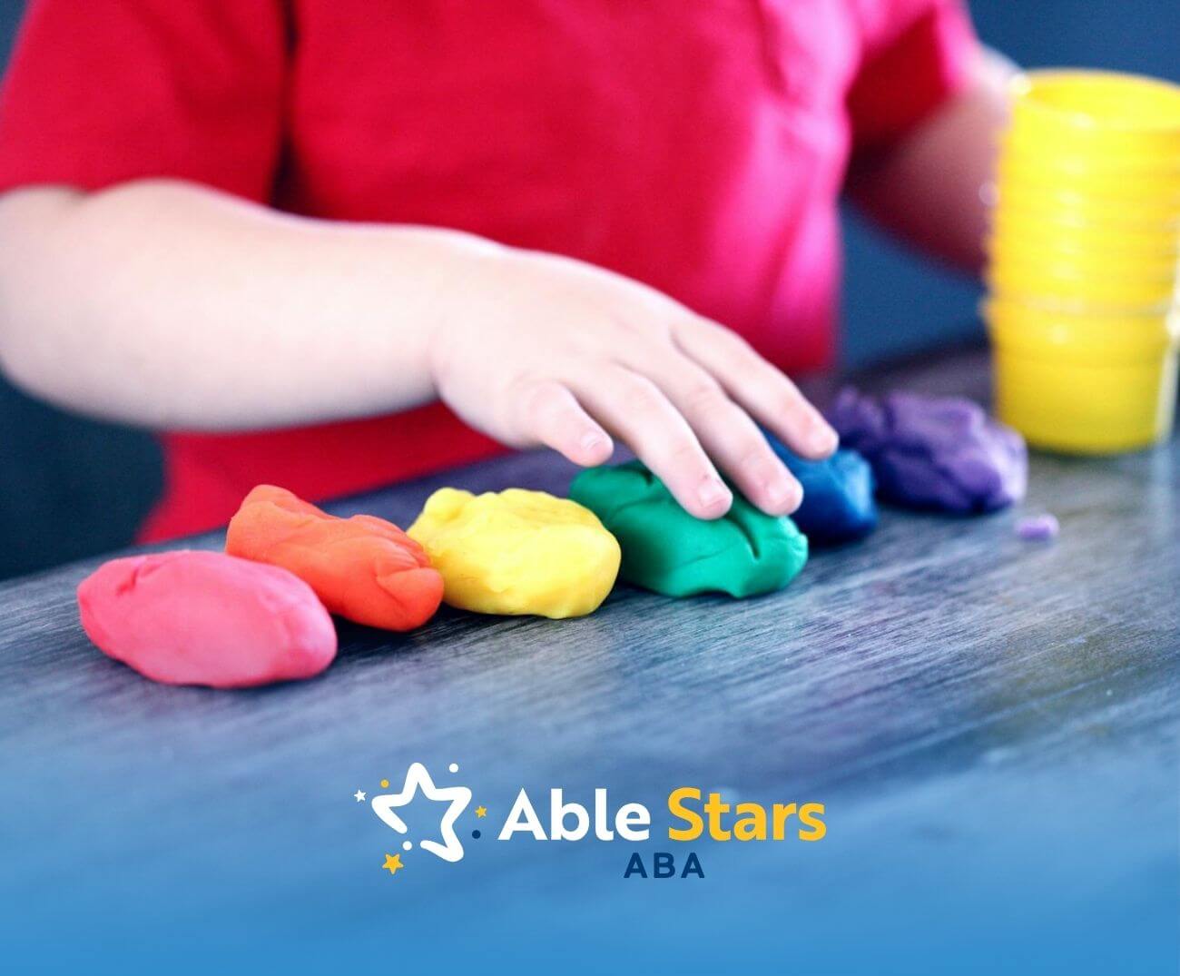 Child in red shirt reaches out to sort rainbow-colored playdough pieces on table, engaging in creative, tactile play.