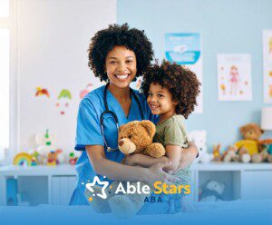 Smiling healthcare worker in blue scrubs hugs a child holding a teddy bear. They stand in a colorful, cheerful room, radiating warmth and care.