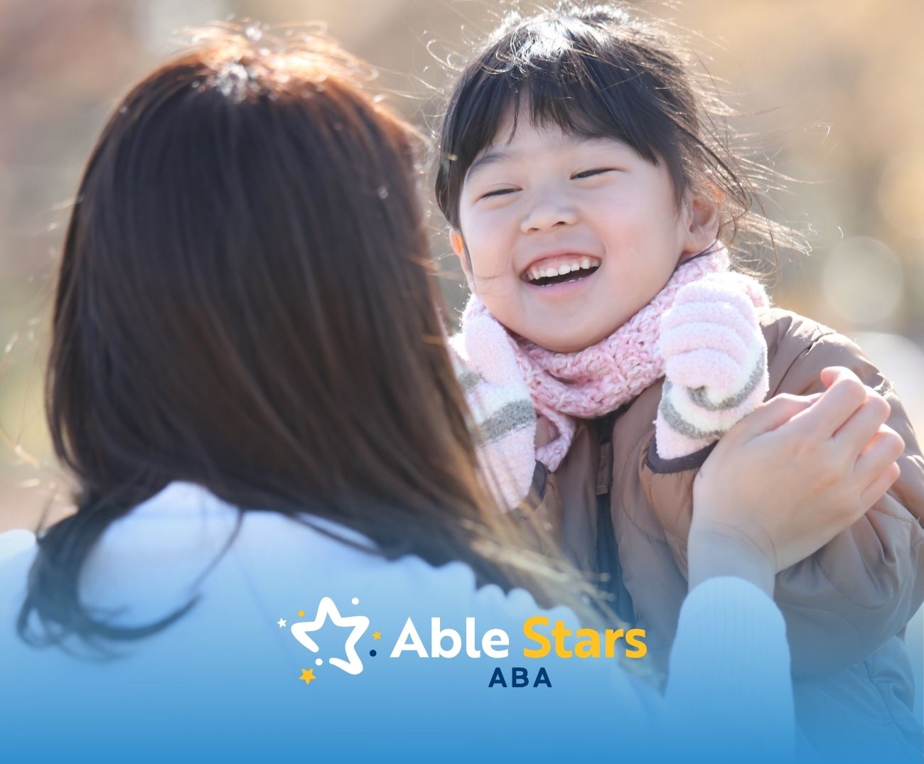 A young girl wearing a scarf smiles joyfully at an adult with long hair, who holds her hands.