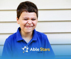 A young boy in a blue polo squints and playfully scrunches his face, standing against a light wooden wall.