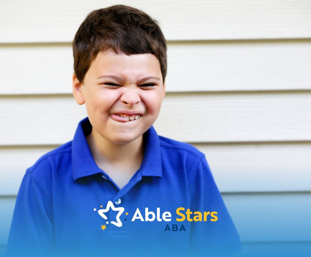 A young boy in a blue polo squints and playfully scrunches his face, standing against a light wooden wall.